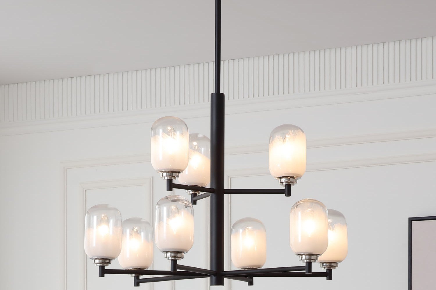 VidaLite Seren 9 Light Chandelier in matte black with milk frost glass shades, lit, hanging against a paneled white wall