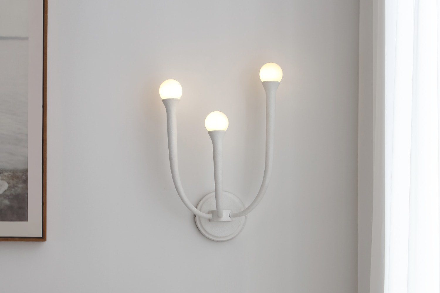 Matte white three light wall sconce lit beside a framed print