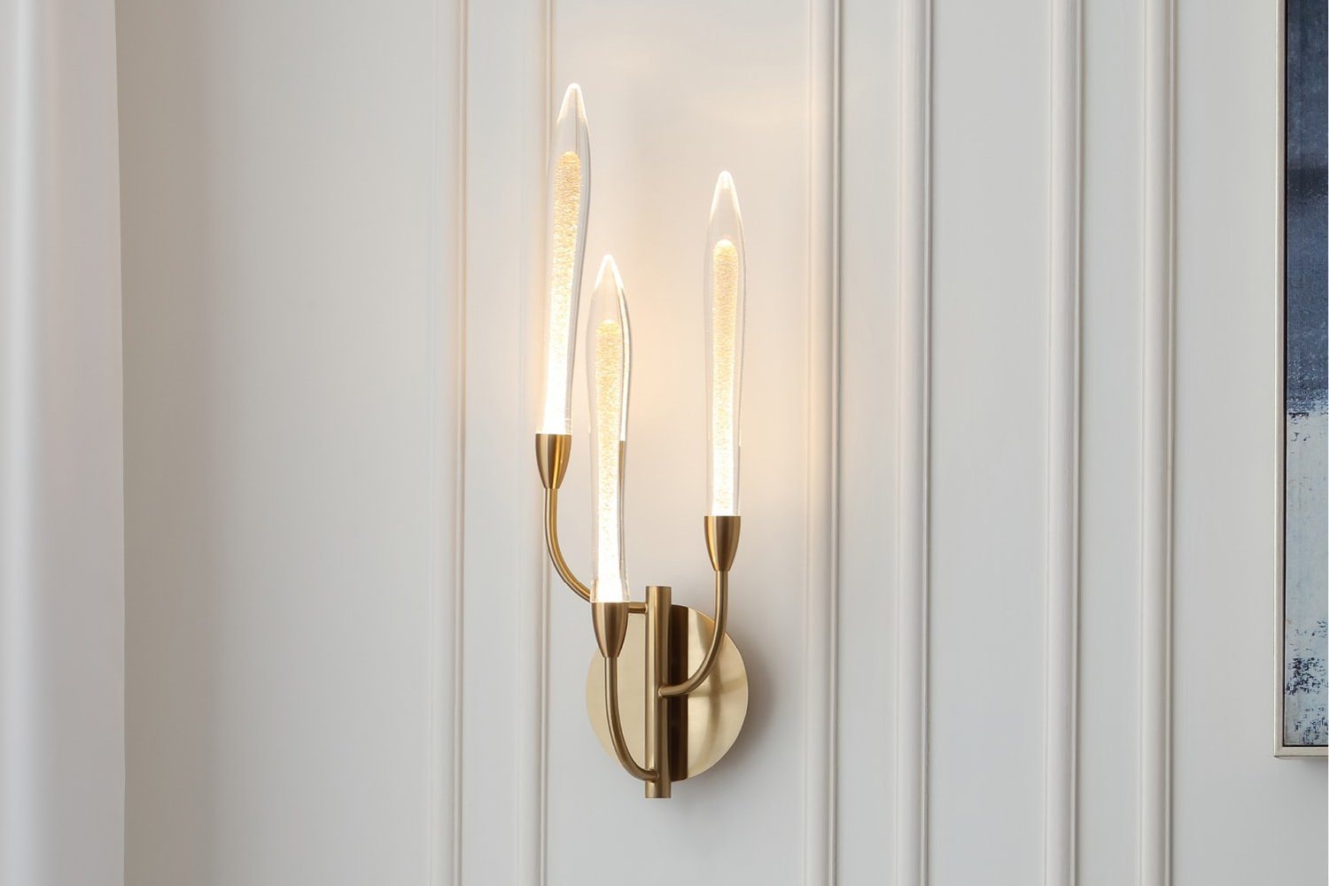 Brass three light wall sconce lit on a white paneled wall next to framed artwork